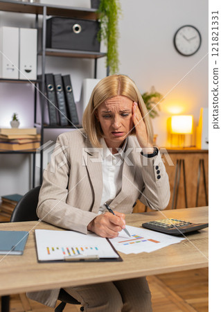 Upset mature businesswoman using calculator feeling worried about financial problem at office desk 121821331