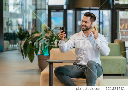 Excited young businessman in headphones listening energetic music on smartphone in office lobby Excited young businessman in headphones listening energetic music on smartphone in office lobby 121821581