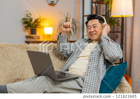 Happy relaxed overjoyed Chinese middle-aged man in wireless headphones listening to music on sofa Happy relaxed overjoyed Chinese middle-aged man in wireless headphones listening to music on sofa 121821605