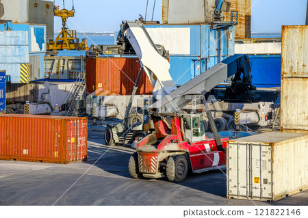 Two reach stackers and cranes transporting large cargo containers at shipping port. Bright sunlight, clear blue sky, stacked containers of various colors, shipping operations of sea cargo port. Two reach stackers and cranes transporting large cargo containers at shipping port. Bright sunlight, clear blue sky, stacked containers of various colors, shipping operations of sea cargo port. 121822146