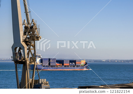 Distant bulk carrier fully loaded with shipping containers leaving harbor and commercial port on sunny day. Ship-to-shore floating crane part in foreground.  121822154