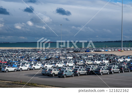 Aerial view of a vast parking lot at seaport cargo terminal. Rows of new vehicles fill the space, creating a colorful mosaic. Cars await shipment to global destinations via nearby Ro-Ro vessels. Scale 121822162