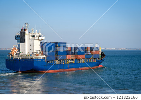 Modern blue bulk carrier loaded with stacked shipping containers navigating harbor near sea port. Global trade, maritime logistics, cargo, international freight. Modern blue bulk carrier loaded with stacked shipping containers navigating harbor near sea port. Global trade, maritime logistics, cargo, international freight. 121822166