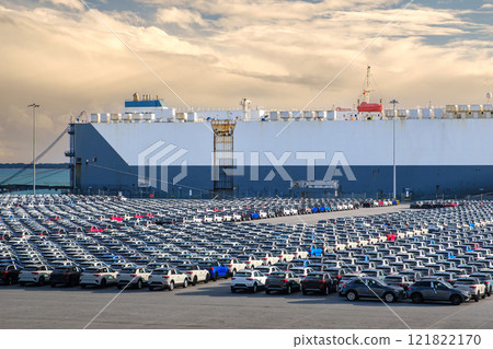 Large vehicle carrier ship known as RO-RO at port on sunny day, large parking area filled with cars waiting loading and transportation to buyers and car dealers. Blue sky and colorful clouds, vehicle 121822170