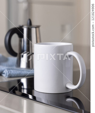 White coffee mug on induction stove against moka pot on blue dish cloth closeup, mockup 121824906