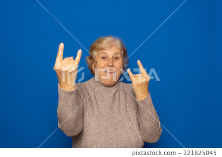 Caucasian woman about 70 years old wearing a dark beige sweater making a rock on sign symbol with hands up smiling broadly, isolated over blue background. Caucasian woman about 70 years old wearing a dark beige sweater making a rock on sign symbol with hands up smiling broadly, isolated over blue background. 121825045
