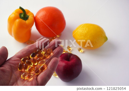 A hand holding several shiny golden capsules is extended in front of a vibrant arrangement of a yellow bell pepper, an orange, a lemon, and a red apple on a white background. 121825054