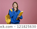 Front view of brunette pretty schoolgirl standing, showing quiet. Beautiful female with earphones and yellow rucksack, folder, looking at camera. Isolated on pink background. 121825630