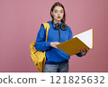 Front view of pretty schoolgirl standing with open mouth, looking at camera. Brunette female holding yellow open folder and rucksack, wearing casual clothes. Concept of youth. 121825632
