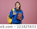 Front view of young pretty student standing, holding present. Beautiful schoolgirl wearing casual clothes, looking at camera, showing super. Isolated on pink studio background. 121825633