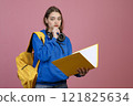 Front view of beautiful schoolgirl standing, holding open folder, book, thinking. Pretty brunette girl looking at camera, touching chin. Isolated on pink studio background. 121825634
