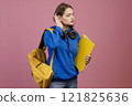 Front view of pretty brunette girl standing, touching ear, listening. Beautiful student with earphones, holding yellow rucksack and folder. Isolated on pink studio background. 121825636