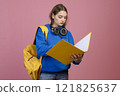 Front view of brunette schoolgirl standing, holding open folder, reading. Pretty young female wearing blue khudi and jeans, earphones, looking down. Concept of modern lifestyle. 121825637