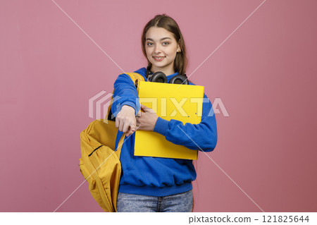 Front view of brunette female brunette standing, holding yellow backpack and rucksack. Pretty schoolgirl wearing blue khudi and jeans, pointing by finger. Concept of youth life. Front view of brunette female brunette standing, holding yellow backpack and rucksack. Pretty schoolgirl wearing blue khudi and jeans, pointing by finger. Concept of youth life. 121825644