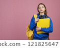 Front view of pretty female with rucksack and earphones standing with closed eyes. Beautiful schoolgirl wearing blue khudi, holding folder. Isolated on pink studio background. 121825647