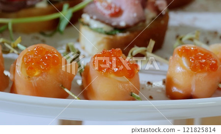 Gourmet appetizer featuring delicate smoked salmon rolls crowned with bright red caviar, elegantly plated with fresh dill beside complementary canapes 121825814