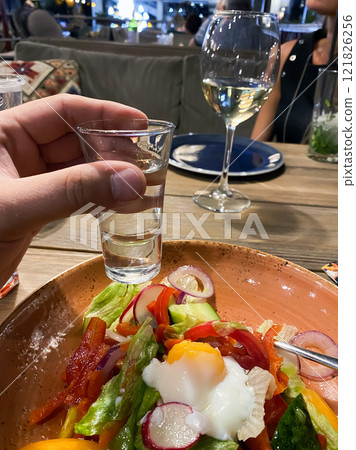 shot glass with alcohol in hand in a restaurant on a background of appetizers and vegetable salad 121826256