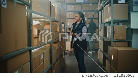 Female Warehouse Associate Scans Cardboard Boxes, Uses Tablet Computer, Takes Parcel for Shipping 121826602