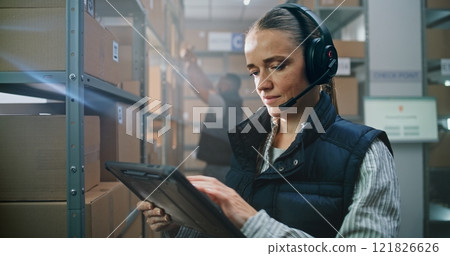 Female Logistics Coordinator Wearing Headset Uses Tablet Computer in Warehouse Facility 121826626