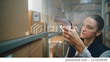 Female Warehouse Associate Scans Cardboard Box, Takes Parcel from Shelf for Shipping Female Warehouse Associate Scans Cardboard Box, Takes Parcel from Shelf for Shipping 121826699