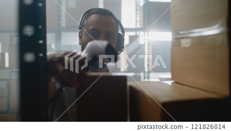 African American Warehouse Associate Talks on Headset, Scans Cardboard Box, Uses Tablet Computer 121826814