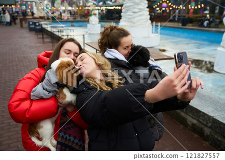 Friends enjoying a playful moment while taking a selfie with their dogs near a festive outdoor area in winter 121827557
