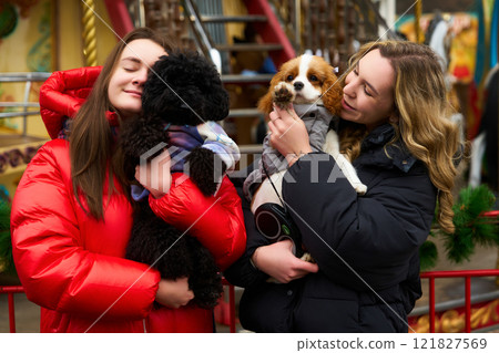 Two friends joyfully holding their small dogs at a festive outdoor carnival during winter Two friends joyfully holding their small dogs at a festive outdoor carnival during winter 121827569