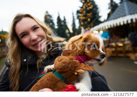 Woman smiles while holding a dog with a festive toy at a holiday market in a winter setting 121827577
