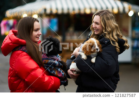 Two friends enjoy a winter day outside while holding small dogs at a festive outdoor market 121827587