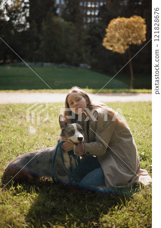 owner walks on lawn in park with corgi dog. beautiful girl with long hair in beige raincoat. animal owner walks on lawn in park with corgi dog. beautiful girl with long hair in beige raincoat. animal 121828676