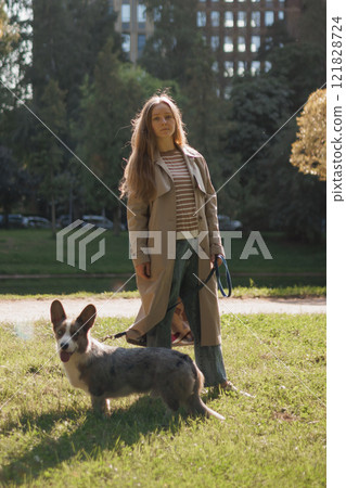 owner walks on lawn in park with corgi dog. beautiful girl with long hair in beige raincoat. animal  owner walks on lawn in park with corgi dog. beautiful girl with long hair in beige raincoat. animal  121828724