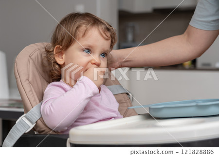 A toddler in a highchair enjoying a snack while being gently supervised at home. A toddler in a highchair enjoying a snack while being gently supervised at home. 121828886