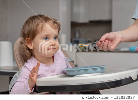 A toddler in a highchair attentively watching a spoon during mealtime in a cozy home. A toddler in a highchair attentively watching a spoon during mealtime in a cozy home. 121828887
