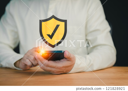 Close-up of a person holding a smartphone with a glowing shield icon featuring a check mark, symbolizing mobile security, data protection, and secure online verification. Close-up of a person holding a smartphone with a glowing shield icon featuring a check mark, symbolizing mobile security, data protection, and secure online verification. 121828921