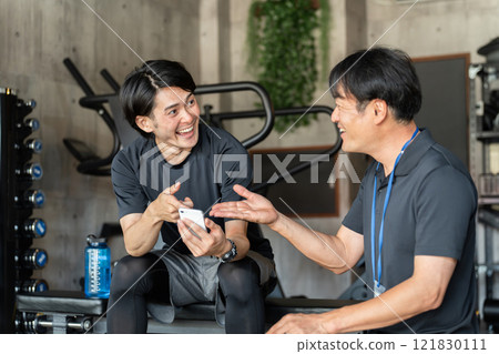 一名男教練使用智慧型手機向健身房/體育館的男性會員通報優惠資訊。 一名男教練使用智慧型手機向健身房/體育館的男性會員通報優惠資訊。 121830111