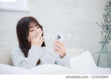 A young woman yawning and operating a smartphone in bed after waking up in the morning 121830904