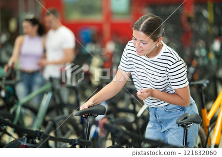 Smiling female cyclist choosing bike with comfortable saddle in store 121832060