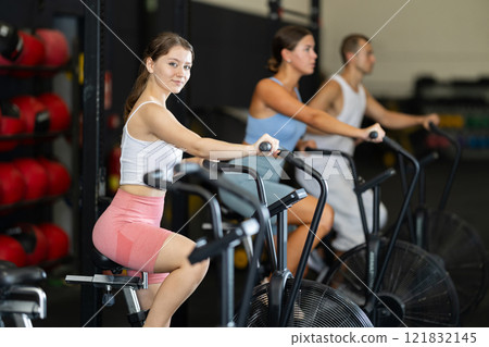 Girl engaged in air bike workout with group of sporty people in exercise room during weight training classes Girl engaged in air bike workout with group of sporty people in exercise room during weight training classes 121832145