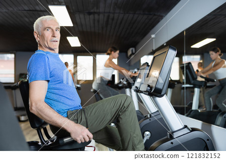 Elderly man, along with other people, is engaged on an bike in gym 121832152