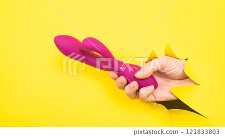Female hand with magenta vibrator sticking out from yellow paper background. Copy space. Female hand with magenta vibrator sticking out from yellow paper background. Copy space. 121833803