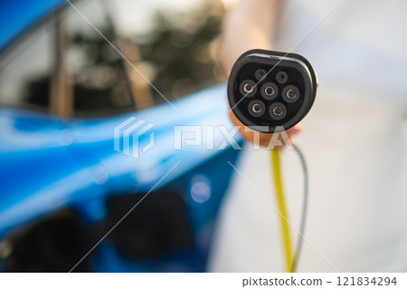 Woman holds a charging connector while charging her electric vehicle at a charging station. 121834294