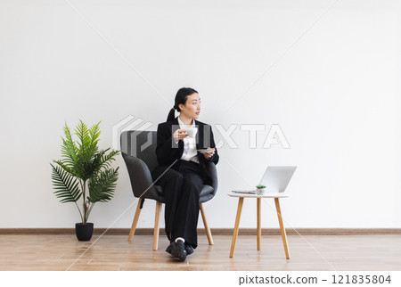 Asian businesswoman seated with coffee in minimalist office setting. Woman in professional suit working on laptop, creating calm business environment. Modern, clean design emphasizes productivity. Asian businesswoman seated with coffee in minimalist office setting. Woman in professional suit working on laptop, creating calm business environment. Modern, clean design emphasizes productivity. 121835804