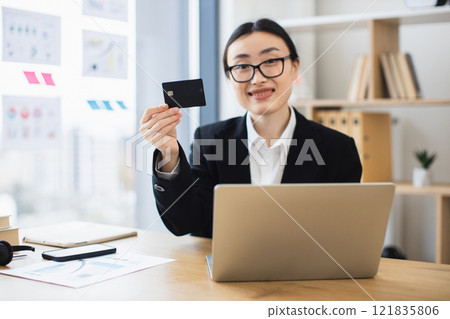 Asian woman in business suit holding credit card at office desk, using laptop for online banking. Smiling confidently, showcasing modern technology and professional success. 121835806