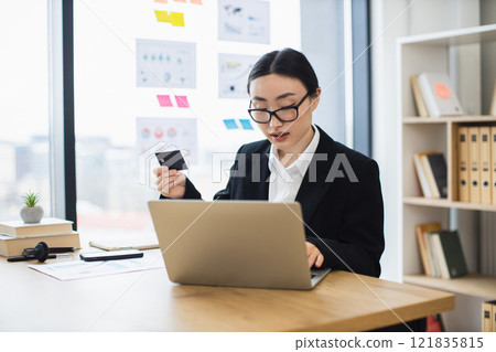 Asian business woman using credit card for online payment at office desk. Young female working on laptop, efficient and focused. Asian business woman using credit card for online payment at office desk. Young female working on laptop, efficient and focused. 121835815