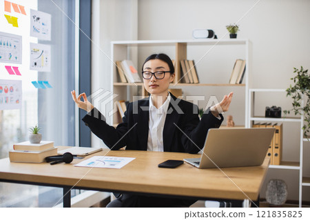 Asian businesswoman meditating at desk, finding balance during office break. Adult female in formal attire, calm atmosphere, bright workspace. 121835825