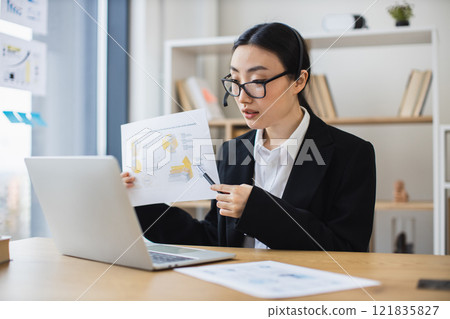 Asian female professional in formal attire conducts online meeting, presenting analysis report on laptop. Modern office setting shows productivity, communication, expertise. 121835827