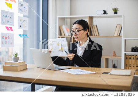 Asian female professional in formal attire conducts online meeting, presenting analysis report on laptop. Modern office setting shows productivity, communication, expertise. Asian female professional in formal attire conducts online meeting, presenting analysis report on laptop. Modern office setting shows productivity, communication, expertise. 121835829
