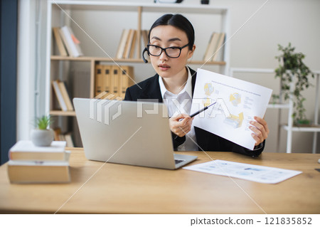 Asian woman in 30s wearing headset engaged in video call, presenting data to colleagues, business setting. Focused and professional atmosphere in modern office space. 121835852