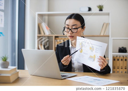 Asian businesswoman wearing headset engages in online video call while reviewing documents at modern office desk. 121835870