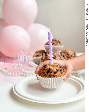 Childs hand holding cupcake with purple candle and chocolate chips next to pink balloons. Birthday concept Childs hand holding cupcake with purple candle and chocolate chips next to pink balloons. Birthday concept 121838767